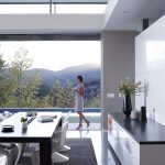 A woman holding a cup walks through a modern, open-concept home with large sliding doors that reveal a scenic mountain view and a poolside patio.